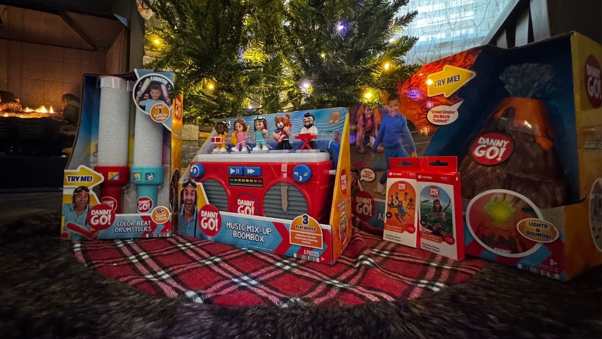 Collage of Danny Go! toys including the Music Mix-Up Boombox with character figures, Color Beat Drumsticks, a dance mat, aviator hat and goggles, and a volcano-shaped speaker, with Danny jumping in the center