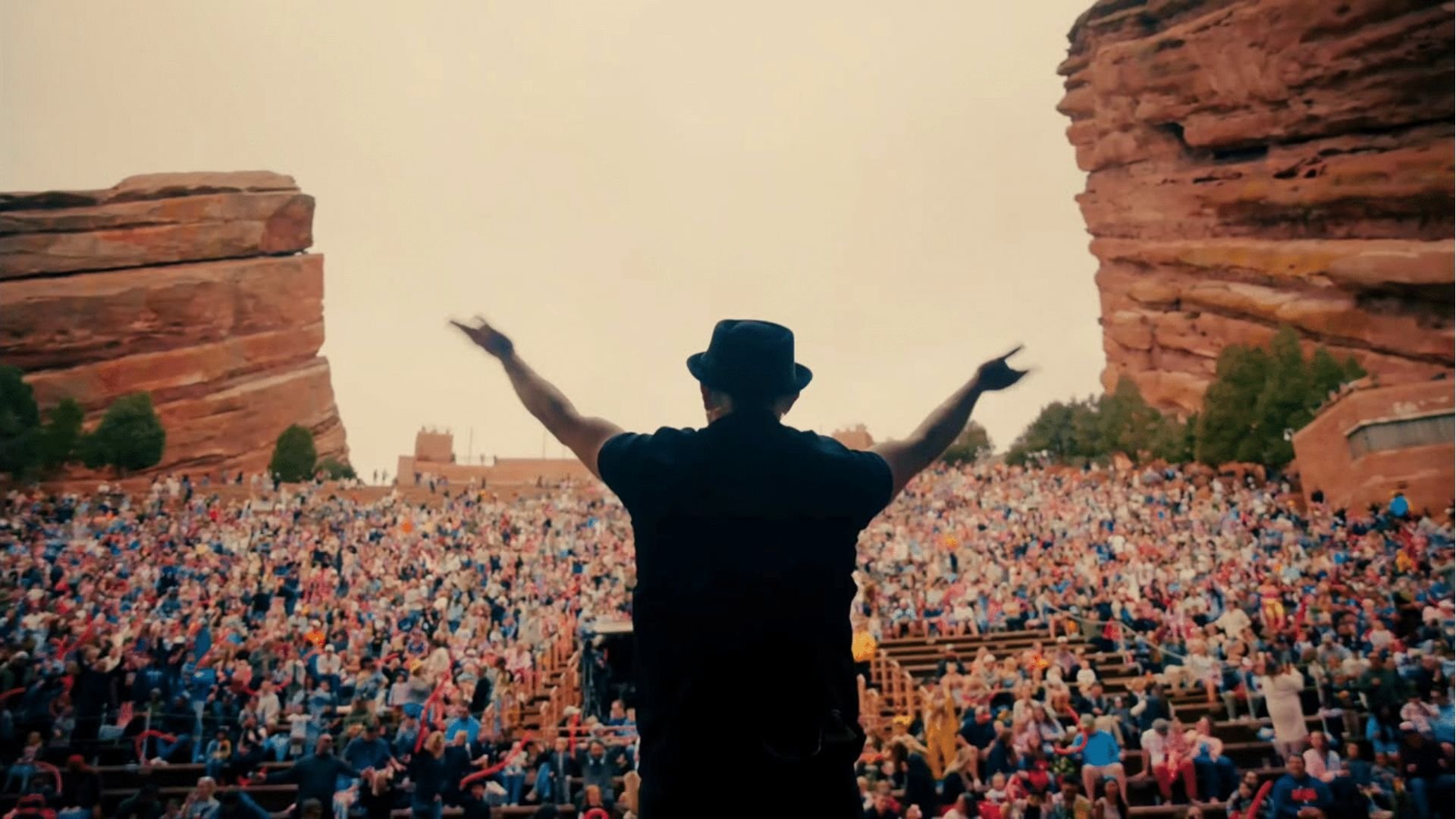 DJ Raphi on stage at Red Rocks Amphitheatre with arms raised, facing a packed crowd between towering red sandstone rocks