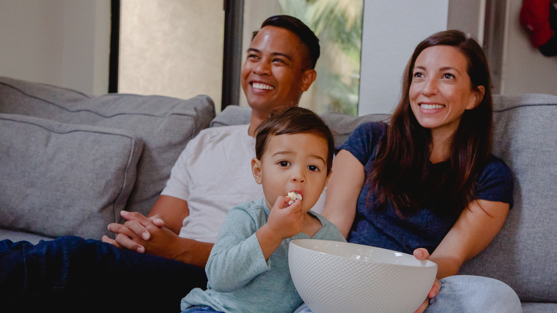 Family relaxing on a couch together while watching faith-based entertainment and sharing popcorn during a family movie night.