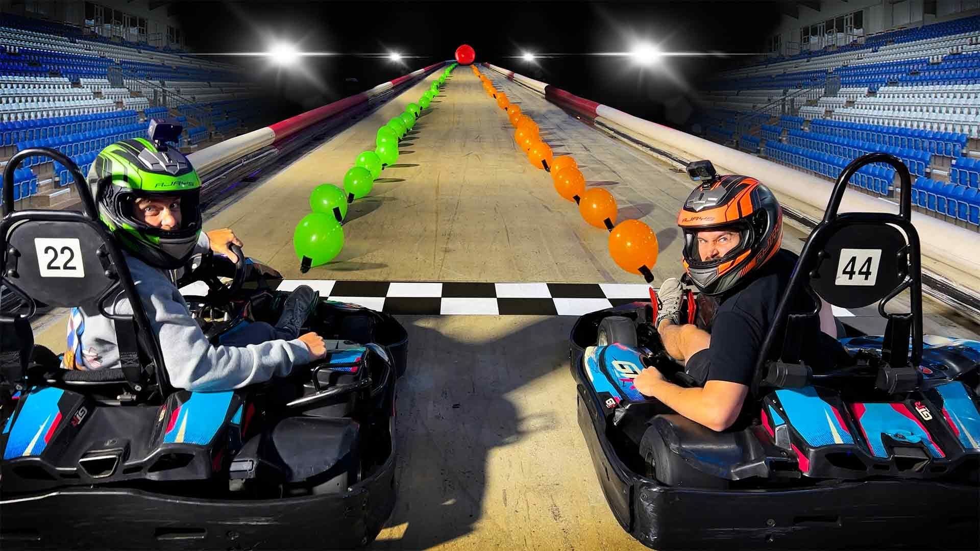 Two people in go-karts, numbers 22 and 44, look back at the camera at the starting line of a track lined with green and orange balloons in the How Ridiculous balloon-pop race.